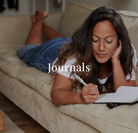 Woman lying on a beige couch, writing in a journal with a pen. Calm home setting with soft lighting and the word "Journals" centered over the image.